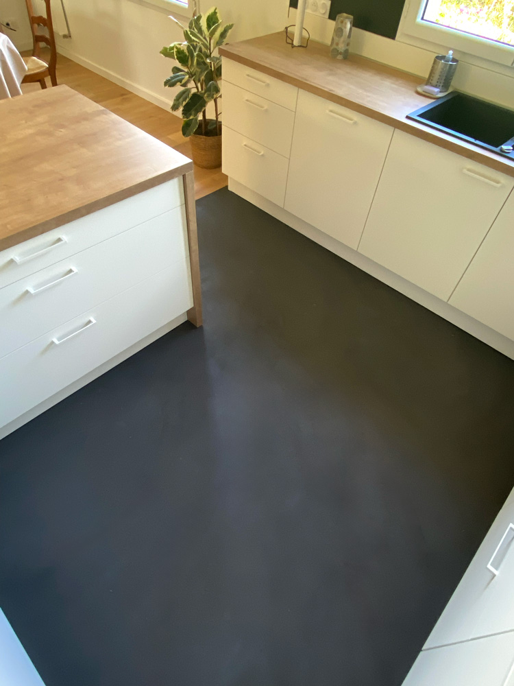 Pose de béton ciré noir mat pour le sol d'une cuisine au mobilier bois et blanc. Le noir apporte contraste et personnalité à la pièce.